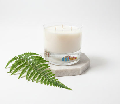 A three-wick soy candle in a clear glass jar sits on a white desk in a bedroom setting, with a potted plant and bed visible in the background.