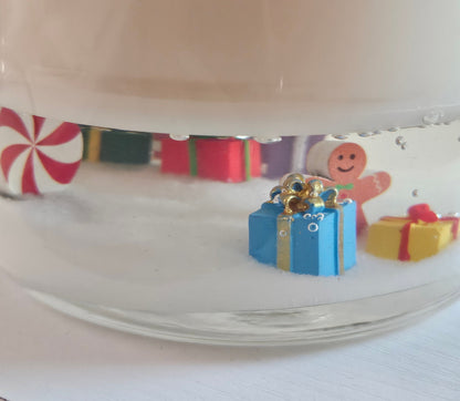 A close-up of a clear glass jar filled with white wax or resin, featuring small colorful holiday decorations inside, including miniature gift boxes, a gingerbread man figure, and a red-and-white peppermint candy illustration.