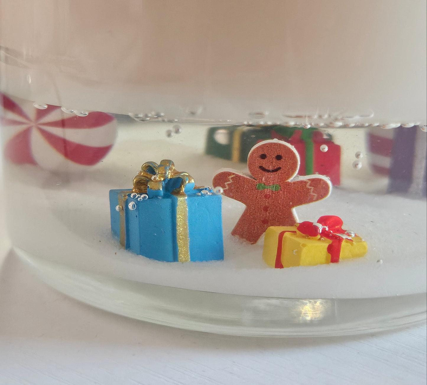 A close-up of a clear glass jar filled with white wax or resin, featuring small holiday decorations inside, including a gingerbread man figure, a blue gift box, and a yellow present, with a blurred pink-and-white striped candy in the background.