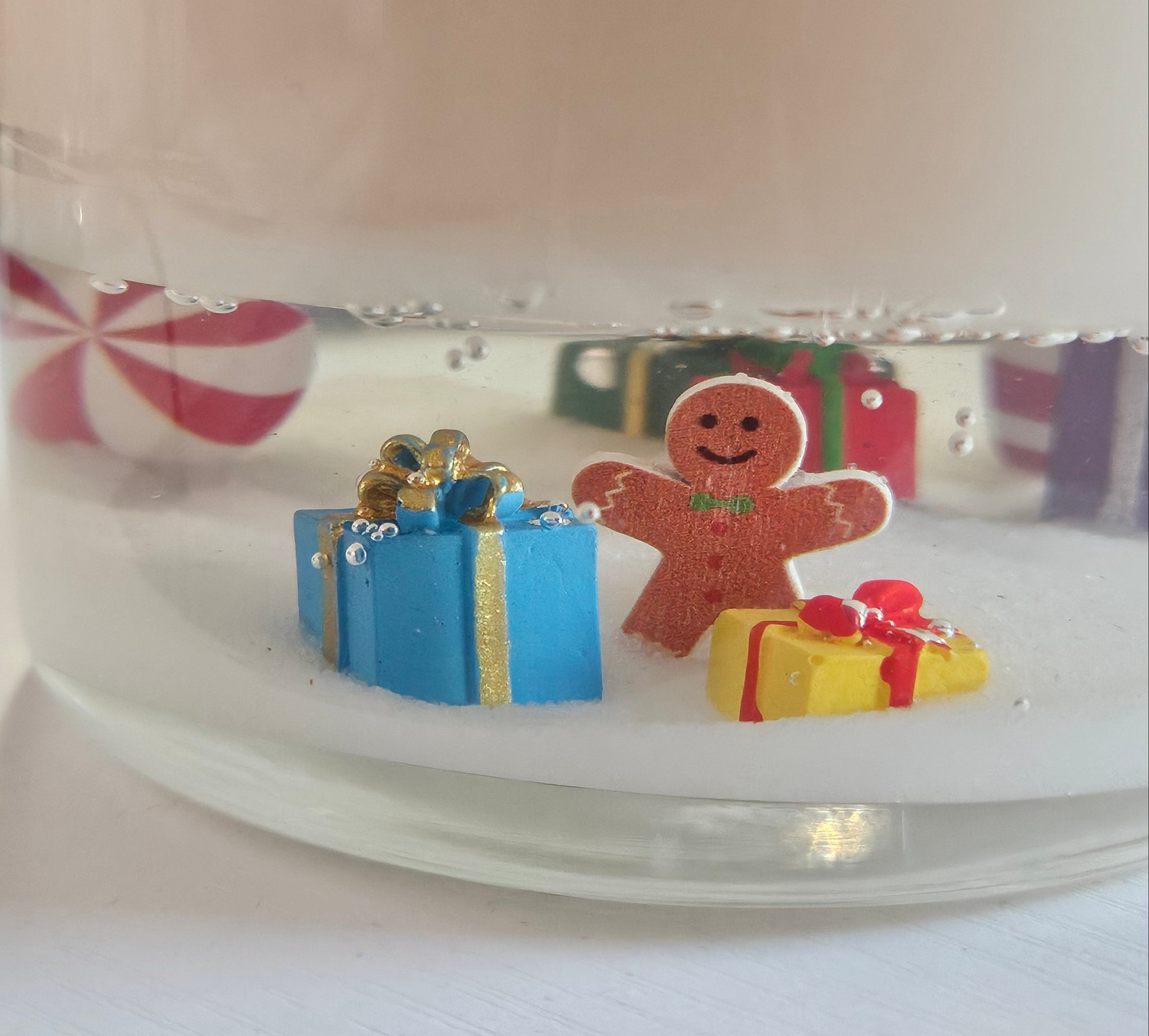A close-up of a clear glass jar filled with white wax or resin, featuring small holiday decorations inside, including a gingerbread man figure, a blue gift box, and a yellow present, with a blurred pink-and-white striped candy in the background.