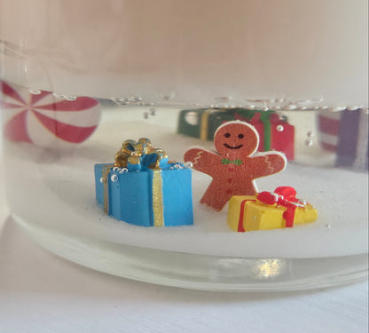A close-up of a clear glass jar filled with white wax or resin, featuring small holiday decorations inside, including a gingerbread man figure, a blue gift box, and a yellow present, with a blurred pink-and-white striped candy in the background.